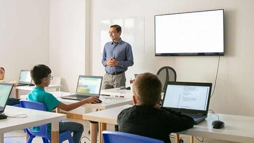 Teacher standing in front of whiteboard with young people using laptops in class