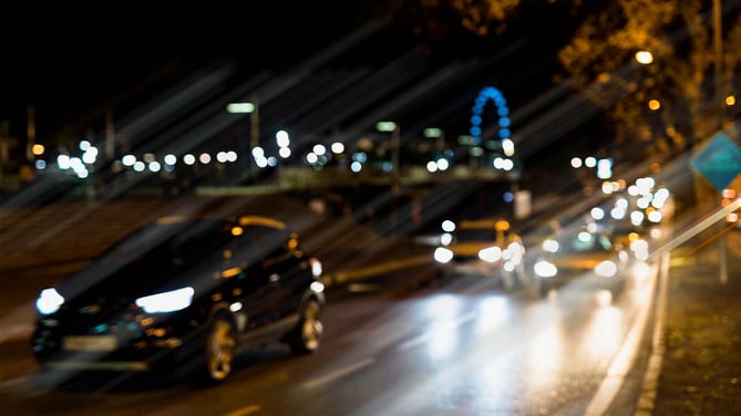 Cars drive down a busy road at night, with their lights on. The car lights and their reflections are bright and streak across the view, the scene is also slightly blurred. This simulates how a person with kerataconus might view this scene