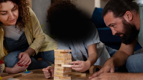 Family playing a Jenga board game. the person in the centre has their face blocked by a large black blob shape, to shows how a person with AMD might visualise the scene.